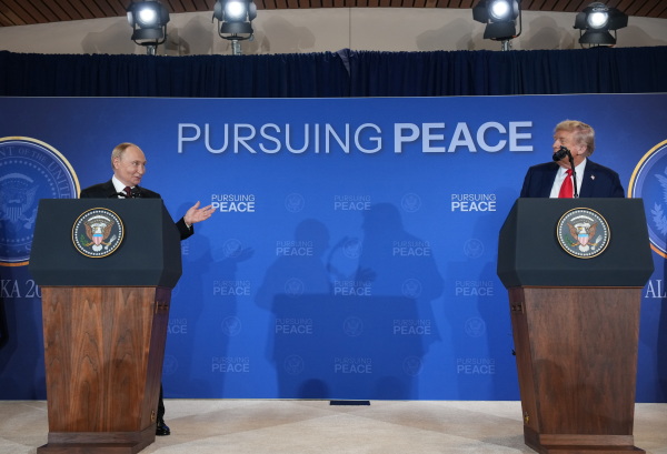 President Trump and Russian President Vladimir Putin are standing outdoors side by side on a blue podium while shaking hands. The front of the podium is displaying in big white letters, "ALASKA 2025." Behind them are fighter jets and Air Force One.