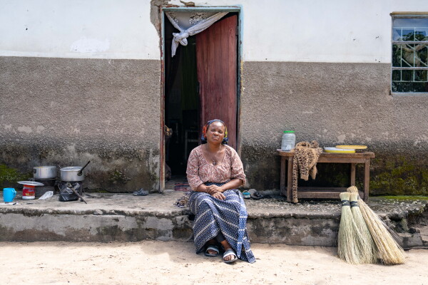 In this photo, Jennifer Mukuka sits on the front stoop of her home. On the left side of the frame, two metal pots rest on burners and on the right side of the frame is a simple wooden table and three bunches of broom bristles. Behind Mukuka is a wooden door to the home, which is partially open.