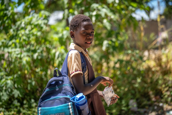 Photographed from the side, Diana is standing outdoors against a backdrop of green foliage. She's wearing a brown jumper dress with a light-brown, short-sleeved shirt underneath. She's carrying a blue backpack on her right shoulder and is holding a foil snack bag.