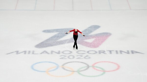  A woman wearing a red shirt and black pants is figure skating. The ice has the words Milano Cortina 2026 on it. 