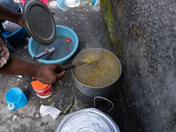 This photo, taken outdoors, shows a metal pot filled with a green stew of pumpkin leaves and groundnuts. In the frame is Jennifer Mukuka's right hand and forearm, stirring the pot with a wooden spoon. On the ground nearby are a light blue plastic basin, a light blue plastic cup and another metal pot.
