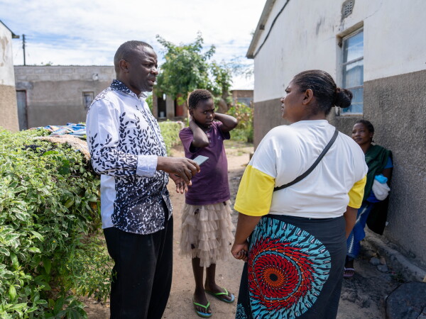 In this photo, Pastor Billy, who's on the left side of the frame, is standing outdoors while talking with Salome Mwale, who's on the right side. Diana is standing behind them, and a woman is seated on the right side of the frame. The side of a one-story structure is on the right, and a row of bushes is on the left.