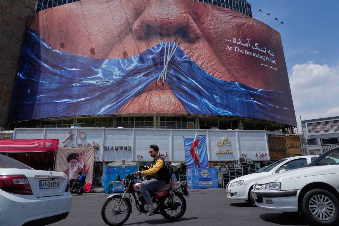 Vehicles drive past a billboard with an illustration showing the Strait of Hormuz and the sewn lips of President Trump in a square in downtown Tehran, Iran, May 2.