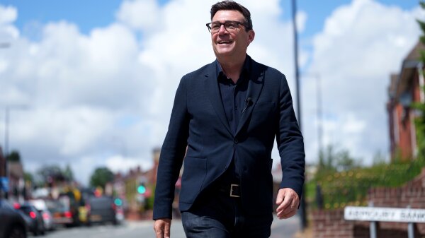 Andy Burnham smiles as he walks. He is wearing an all-dark outfit that features a dress coat.