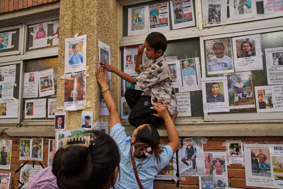 A young boy is helped by a women to put up a photograph of her missing relative on a wall alongside photographs of missing and deceased people displayed outside a government hospital in Kathmandu, Nepal, Sunday, Aug. 30, 2026.