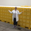 Wearing a white lab coat, dark pants and sneakers, the American Egg Board's Emily Metz stands in front of a multitude of yellow stacked trays that are filled with eggs. She is smiling and is gesturing with her arms stretched out from her sides.