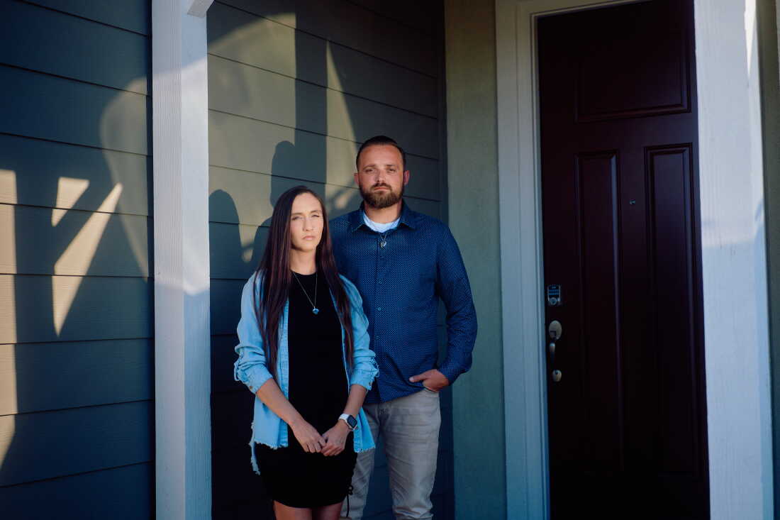 Kaitlyn Morrow and Jeremy Habelitz stand outside of their home in southern California. She is wearing a denim shirt over a black dress and he is wearing a navy button down shirt with khakis.