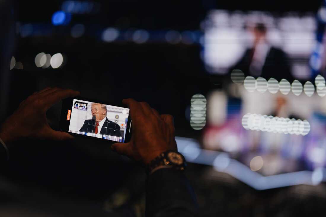This photo shows a pair of hands holding a phone in horizontal position to record Republican presidential candidate Donald Trump speaking on the final night of the 2024 Republican National Convention. The phone's screen shows Trump wearing a suit and red tie while speaking into a microphone. The person holding the phone has a watch on his right wrist, and the photo's background is dark and blurry.