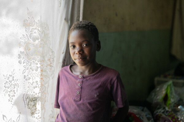 In this photo, Diana stands next to a window inside her home. She has short dark hair and is wearing a purple short-sleeved shirt. The window has a white lace curtain in front of it.