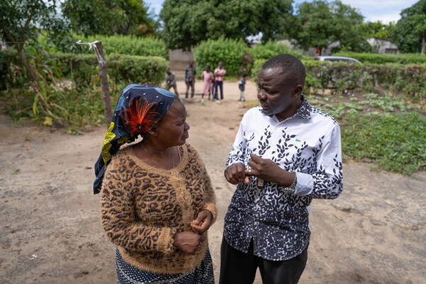 In this photo taken outdoors, Pastor Billy is standing on the right side of the frame while speaking with Jennifer Mukuka, who's on the left side of the frame. Five children are standing in the background at a distance.