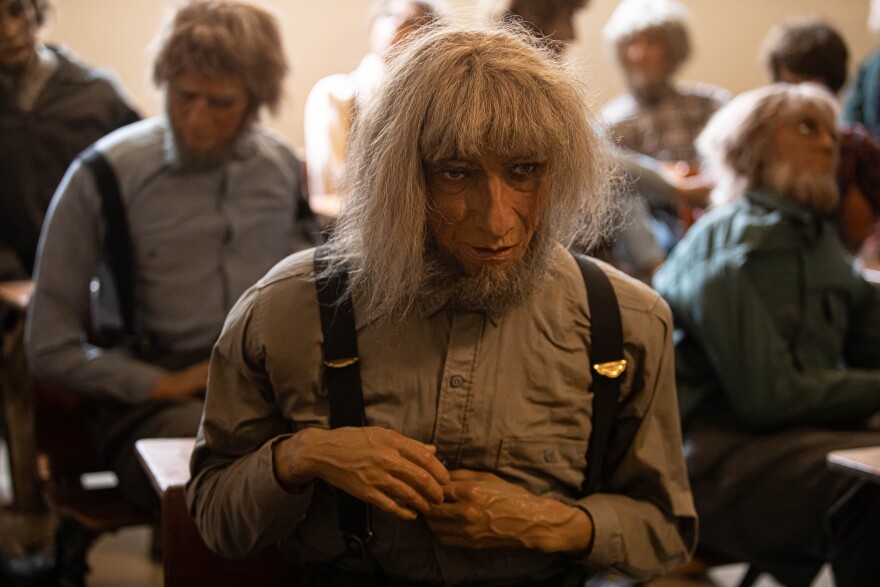 A wax figure of an Amish man with long grey hair and wearing overalls.