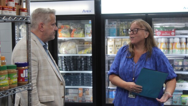 Julie Bonar wears a purple shirt and shows Sorensen the food pantry full of peanut butter, milk, frozen and fresh foods. They stand in front of a refrigerator. 