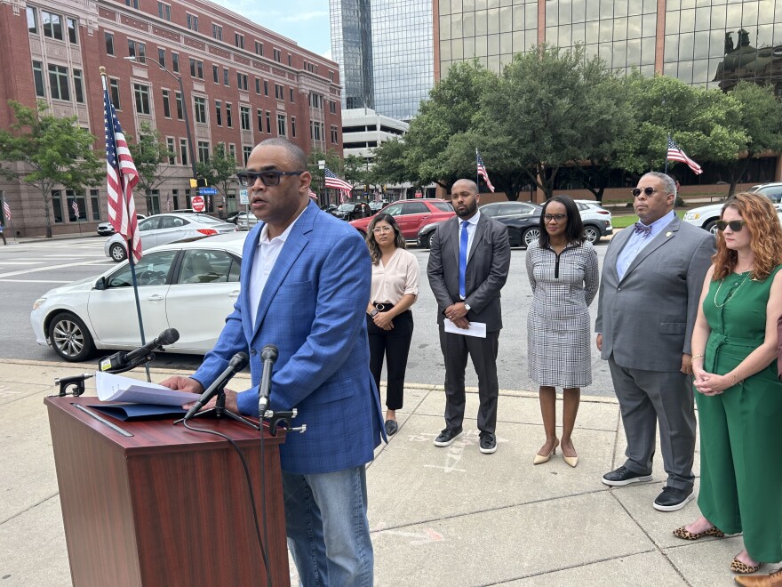 A photo of U.S. Congressman Marc Veasey, a Black man wearing jeans and a blue checkered blazer, speaks at a podium covered in microphones on a downtown street in Fort Worth. Other elected officials line up behind him.