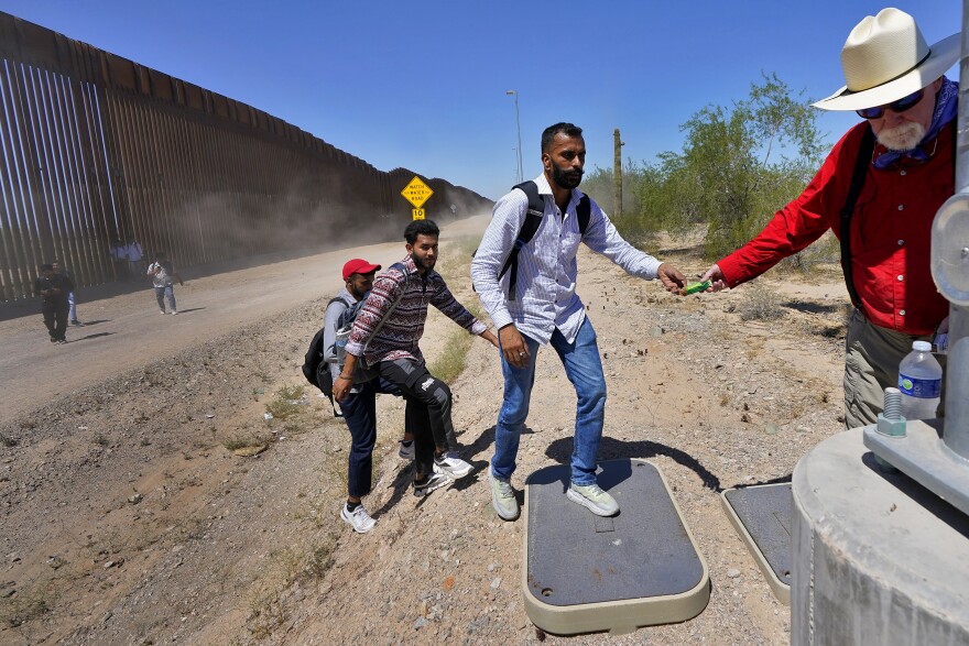 Retired schoolteacher Tom Wingo of Samaritans Without Borders, right, gives snacks and bottles of water to a group of migrants claiming to be from India, who just crossed the border wall, Tuesday, Aug. 29, 2023, in Organ Pipe Cactus National Monument near Lukeville, Ariz. U.S.-Mexico border.