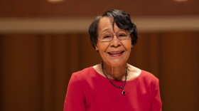 An older woman wearing glasses, a red dress and a dark necklace smiles.