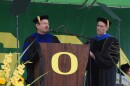 A person speaking at a podium. The person is dressed in academic regalia. The podium is adorned with a large O. A person is standing next to the podium, also dressed in academic regalia.