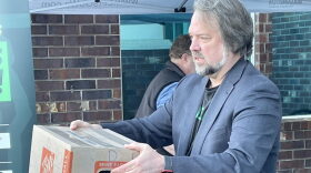 A white man with graying hair and beard in a dark grey blazer holds a cardboard box.