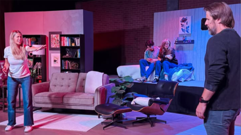 On a theatre stage, a woman with long blonde hair reaches out a hand toward a man with his back to the camera. Between them are two teenagers that sit next to each other on a bed. 