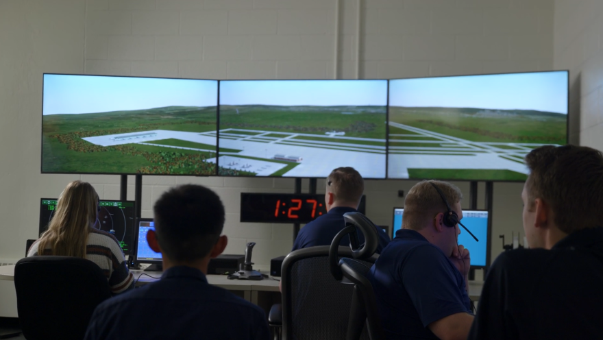 Five people work on computers with three large screens above them. 