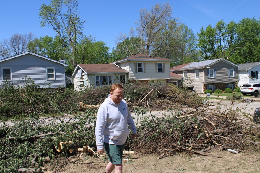 Man in a gray hoodie and green shorts walks away from a line of damaged tree limbs and other debris lined up along a residential sidewalk