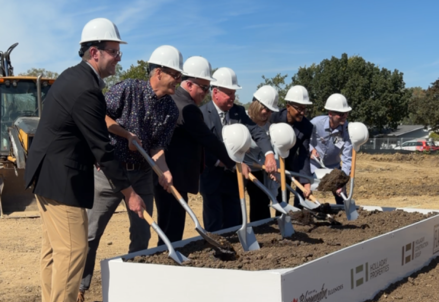 Seven people wearing white construction hats and holding shovels stand in front of a raised box of soil