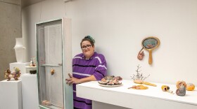 A woman wearing a purple striped shirt stands beside a ceramic installation at SIU’s Sharp Museum, with various colorful ceramic artworks displayed on pedestals and shelves.