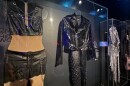 Two stage outfits hang side by side in a glass museum case: a black leather vest and skirt with chain lacing, and a black leather jacket with metal-studded pants. On the right, a silver and denim stage outfit hangs in another glass museum case.