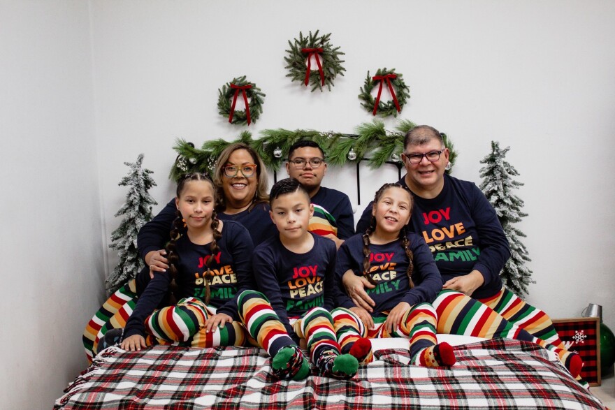 A family Christmas portrait. A mom, dad, and four kids of varying ages sit on a bed with a plaid flannel blanket, surrounded by Christmas decor. They all wear shirts that say "JOY, LOVE, PEACE, FAMILY."