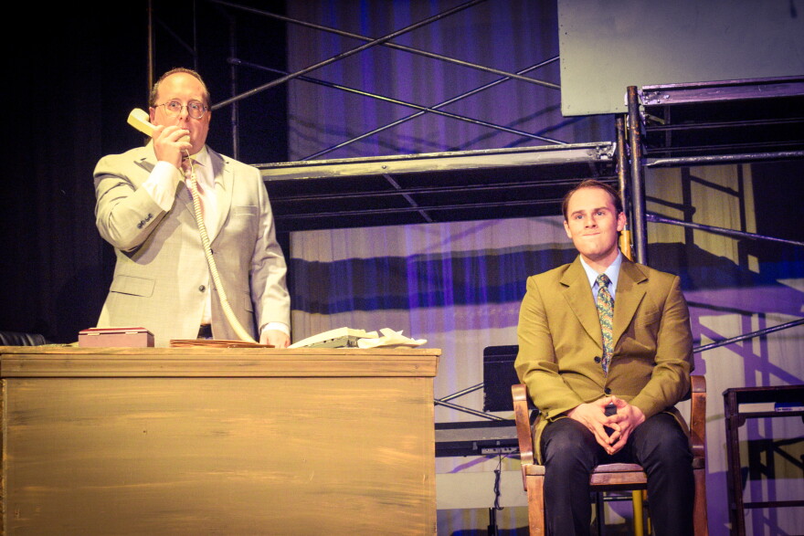 Two men in suits on stage; one stands behind a desk holding a phone to his ear, while the other sits on a chair, hands clasped, smiling slightly. Scaffolding and stage props are visible in the background.