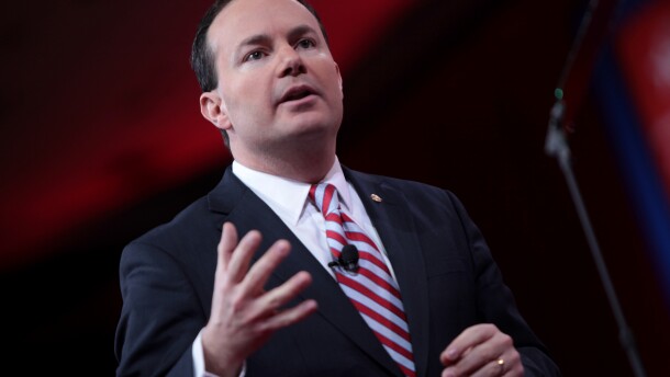 A man in a suit and red and blue striped tie motions at the camera.