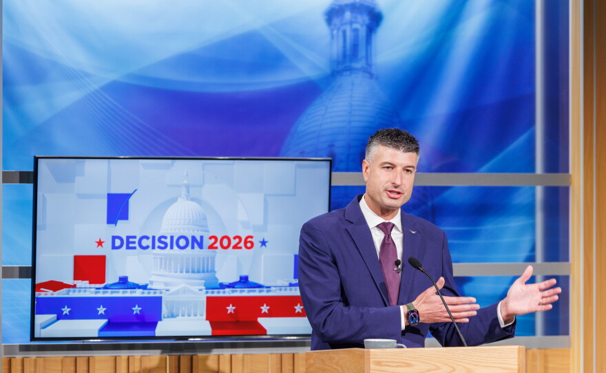 A man in a suit speaks at a podium in front of a TV that reads "Decision 2026"