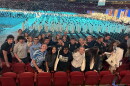 Several dozen high school students pose for a photo in an indoor arena as a gathering of men in white robes walk in a procession at a Catholic church gathering.