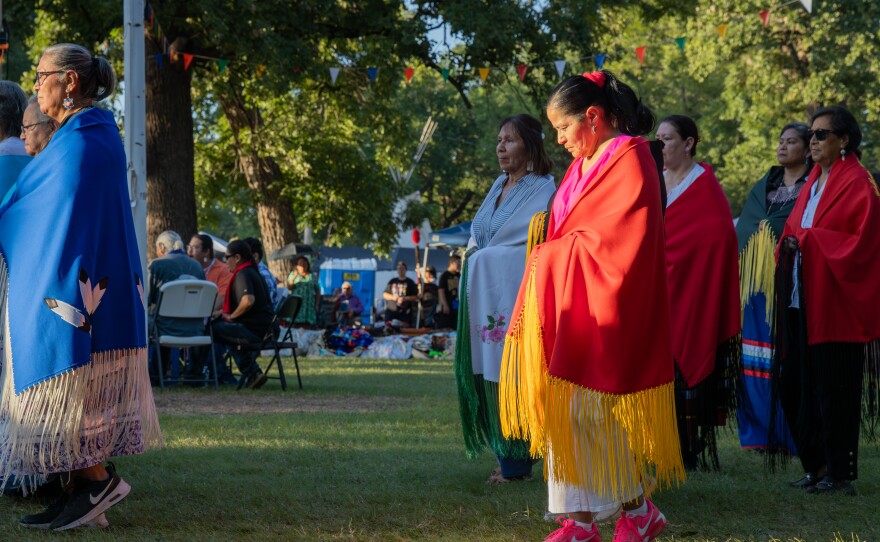 Women wearing colorful robes stand facing the sun with visitors seated in the background at the 72nd annual Comanche Nation Homecoming Powwow on July 19, 2025, in southwest Oklahoma.