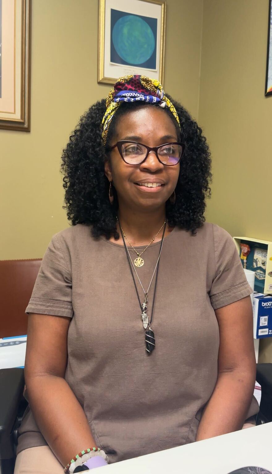 Woman with dark hair and a headband in brown shirt sits in front of sage colored wall while smiling.