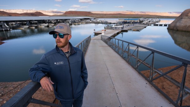 A man wearing a ball cap and jacket stands on a boat ramp in front of a large body of water.