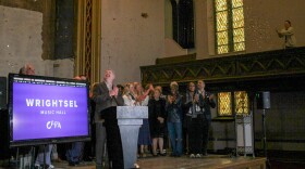People on a stage clap as confetti flutters through the air in an old church. A tall, thin stained glass window glows in one corner. A TV screen displays the name "Wrightsel Music Hall / CAPA" 