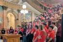 Teens in matching red shirts stand behind a woman who is speaking at a podium. 