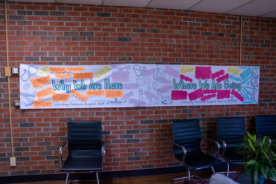 A banner drapes a brick wall inside Newman Memorial Hospital, featuring staff contributions about why they work there and where they hope to go. One quote said, "I work here for the future of rural maternal healthcare that will not be built by one grand moment, but by thousands of small moments of compassion, service, and love."