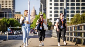 Three women smile while walking on a sidewalk over a bridge in a big city with a capitol dome out of focus in the distance behind them. 