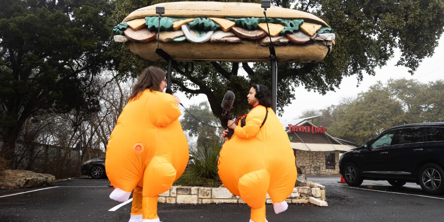 Two people dressed in turkey costumes stand in front of a giant fake sandwich.