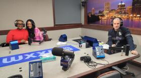 Three people wearing headphones sit at a table in a radio talk studio: a woman at left has short blonde hair and is wearing glasses and a red turtleneck; a woman at center has long dark hair and is wearing a pink tie-dyed sweatshirt; a man at right has short dark hair and is wearing a navy sweater with a wide grey stripe across the chest and sleeves.