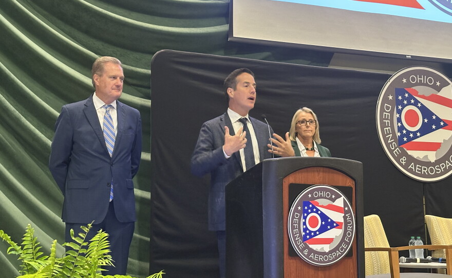A man stands at a podium and speaking, with another man and a woman behind him.