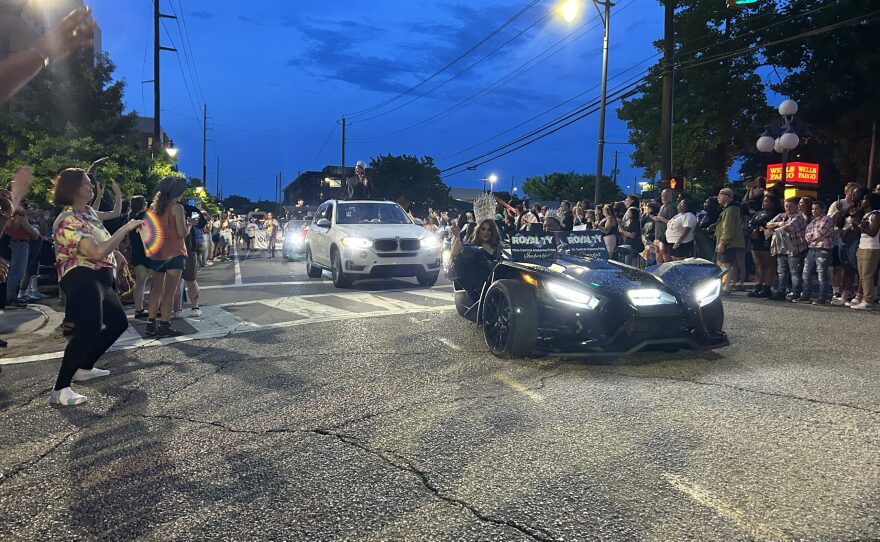A drag queen tosses beads out of a luxury car in the parade while blowing kisses to the crowd.