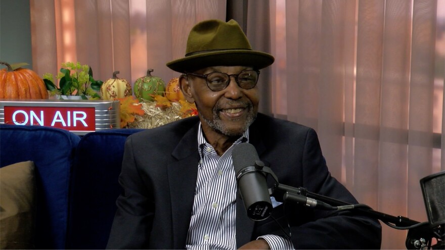 A Black man with round glasses and a gray gotee sits on a dark blue armchair with a microphone stand near his face. He smiles while wearing a dark sport coat, striped button-up shirt and a forrest green hat.