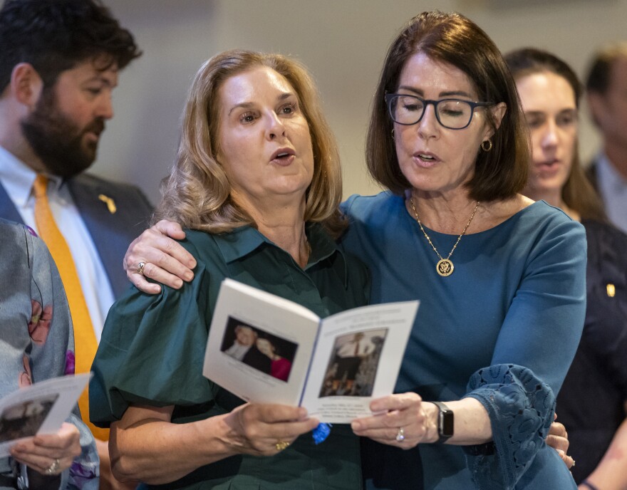 A group of people sing. Two women are in the center of the photo, both holding onto a program. The woman on the left is looking up, the one on the right wears glasses and is looking at the program. She has her arm around the other woman.