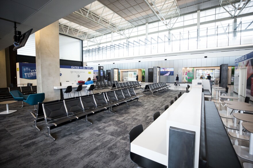 The waiting area at a gate in ABIA. Lots of empty gray seats. High ceilings and windows bring in natural light.