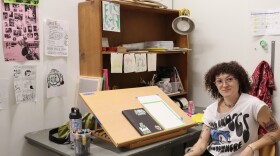 A cartoonist sits at her desk in a studio filled with drawings, posters, markers and sketching supplies.