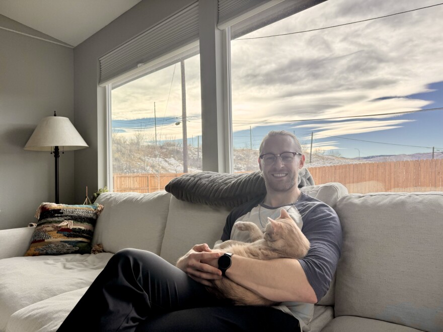 A man sits holding a cat on a grey living room couch in front of a window with a blue sky.