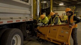 Three men in hard hats and hi-viz jackets hold a plow while one hammers at its attachment to a big truck.