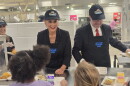 Michigan Governor Gretchen Whitmer and state Senator Sean McCann serve school meals to children at a cafeteria, wearing matching “Champion of the Tray” caps and “Nutrition’s Game One” aprons.